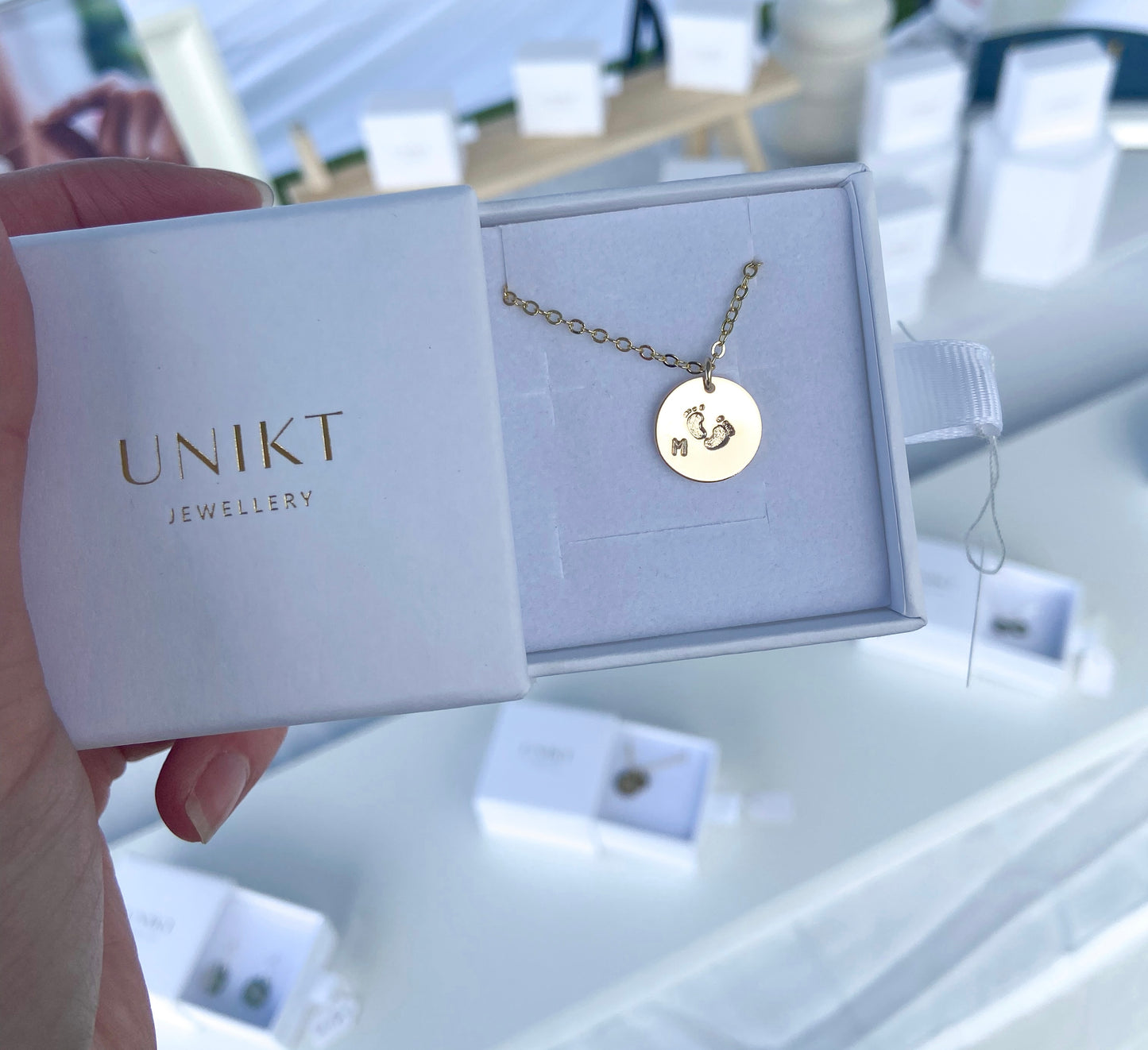 Hand holding a gold footprints necklace with the letter M stamped on it.