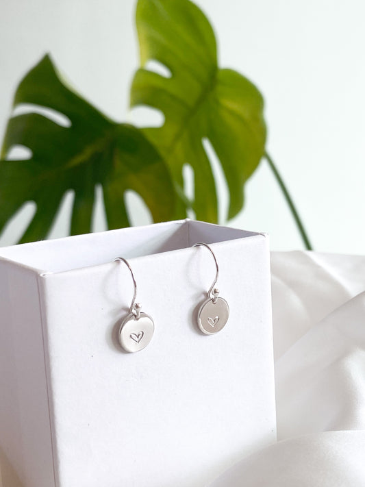 Silver heart earrings on white box with plant in background.